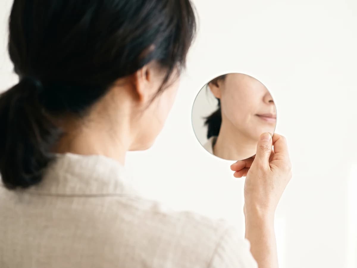 뒤에서 본 여성이 손거울로 얼굴선을 살피고, 거울에 처지기 시작한 볼과 턱선이 비치는 사진
