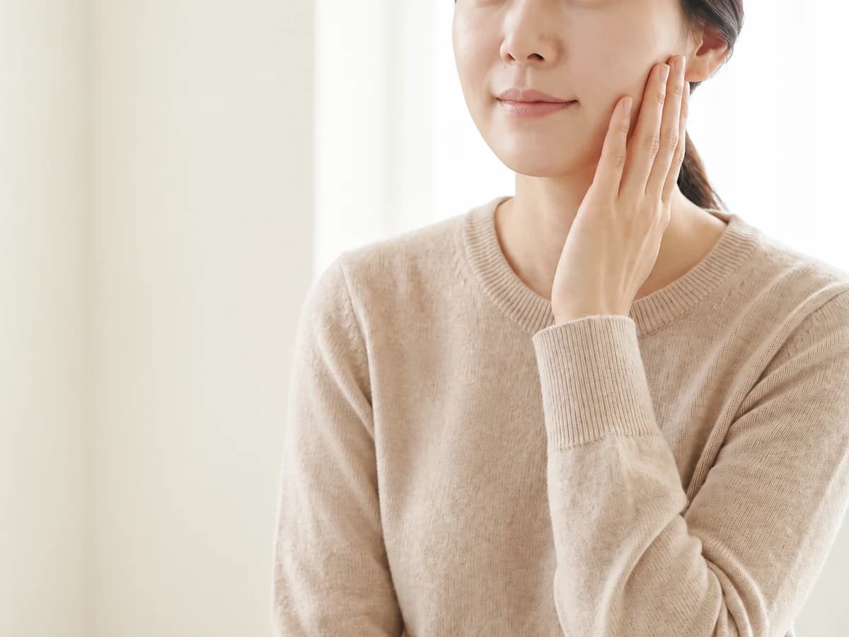 베이지 니트를 입은 여성이 한 손을 볼에 가볍게 얹은 사진